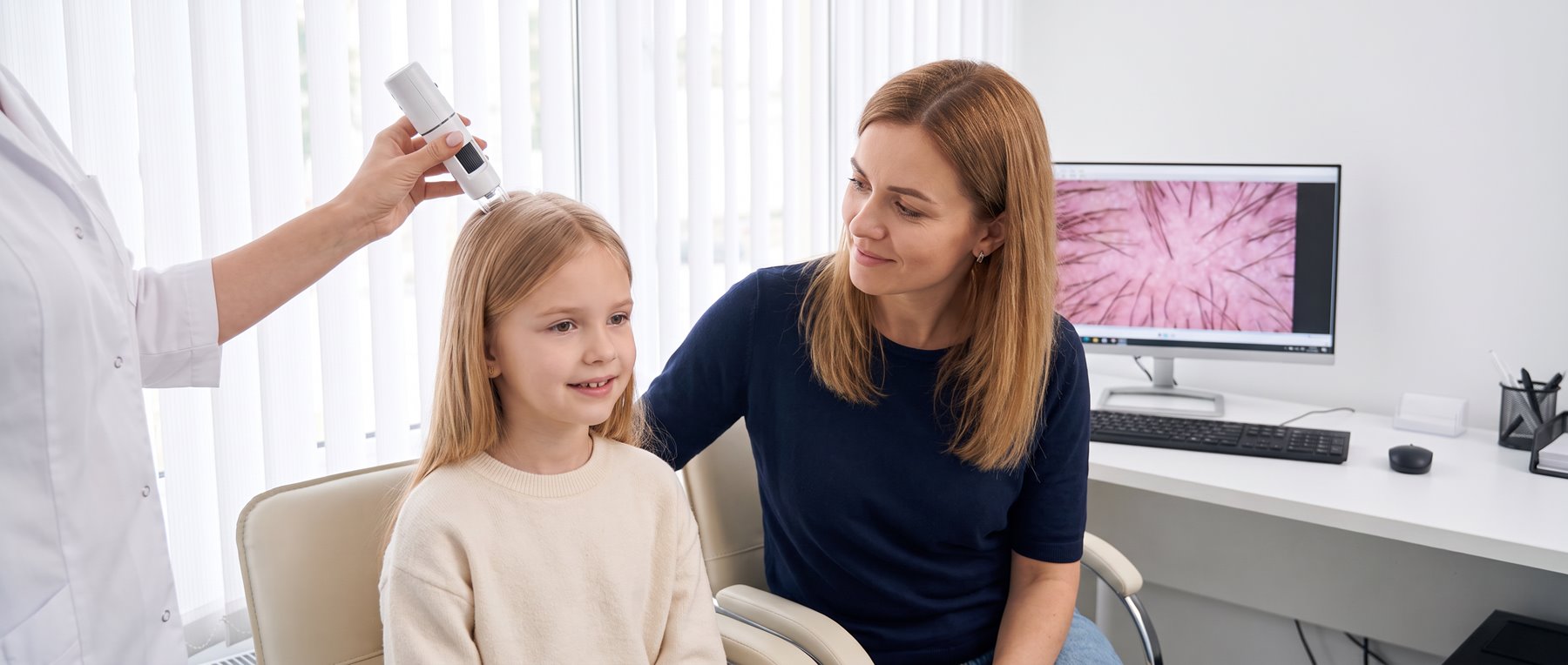 A trichologist examining a young girl’s scalp with a digital microscope while her mother sits beside her, with the magnified view on screen