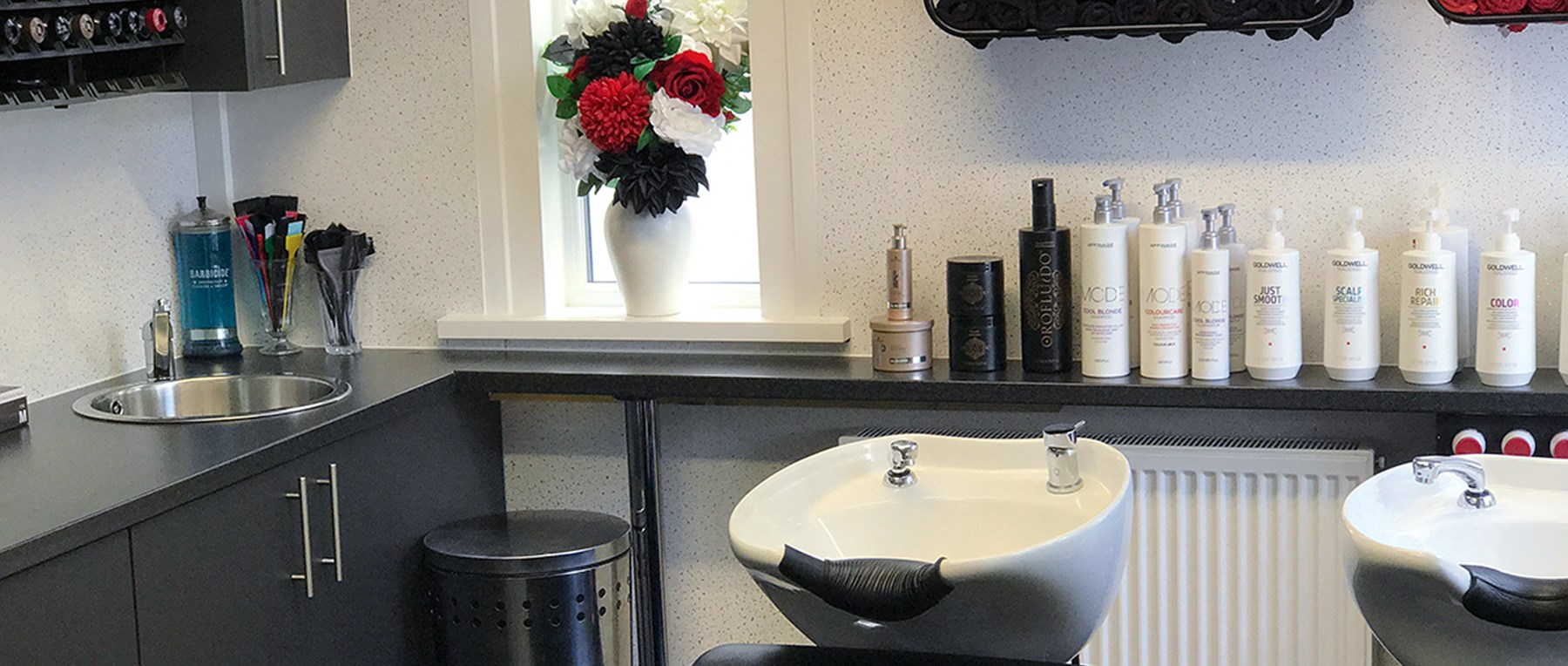 The backwash area at our Plymstock salon: two basins, hair products on the shelf and a vase of red, white and black flowers