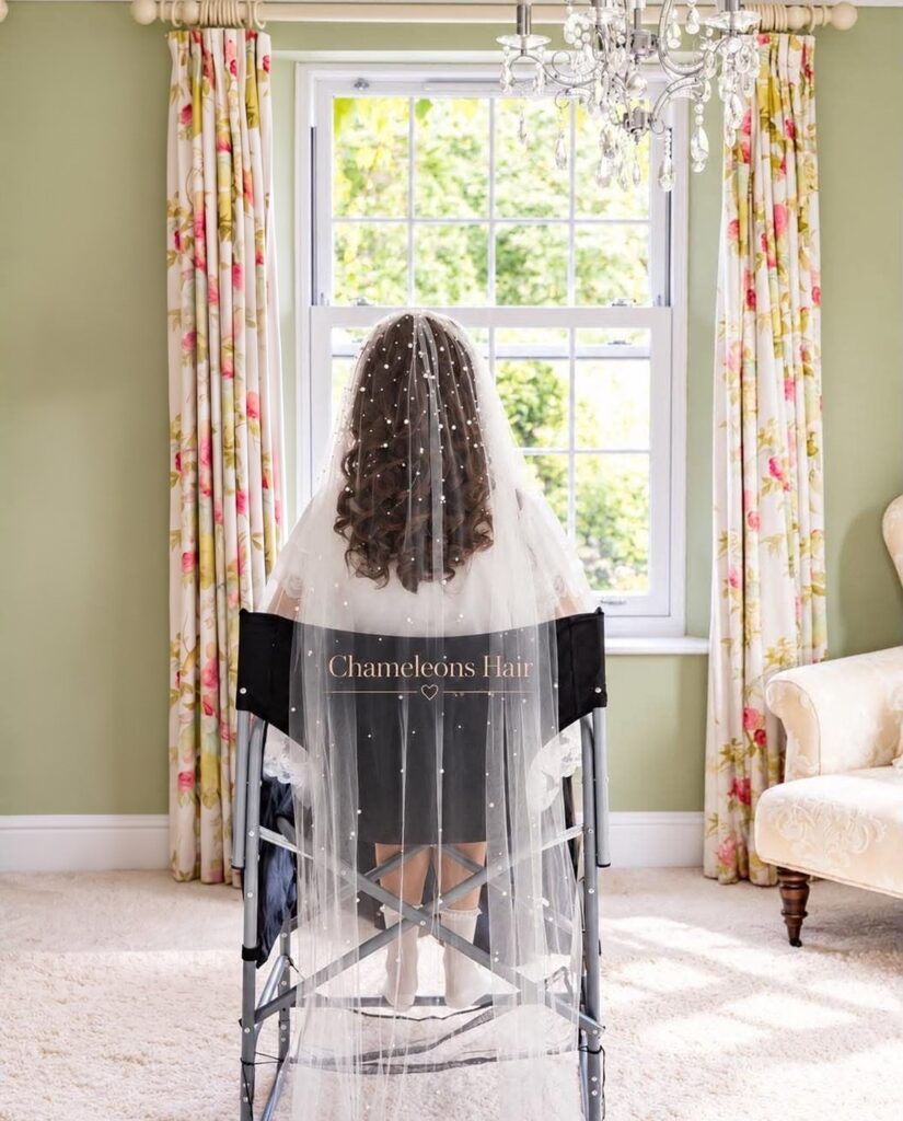 Long dark curls under a pearl-studded veil, seated in a Chameleons Hair chair by a sunny window