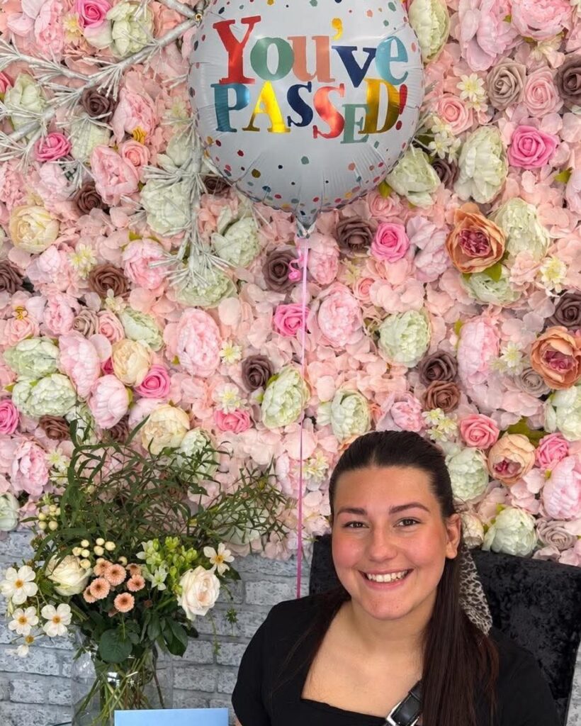 Rebecca smiling in front of a flower wall with a “You’ve passed!” balloon